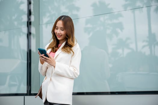 Asian businesswoman typing text on mobile phone standing outdoors street front building near office, Portrait successful business woman smiling holding smartphone using app chat online in morning