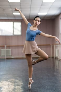 Graceful Asian ballerina in a blue bodysuit and a beige skirt is rehearsing in a dance class