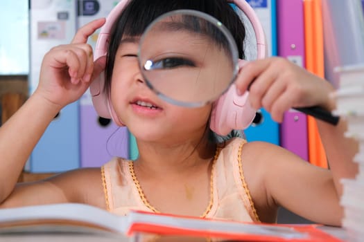 Cheerful little girl in headphones reading a book with a magnifying glass sitting at the table in her room at home.