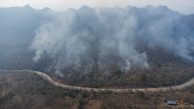 Aerial drone view of a wildfire burning through a forest area, fills the sky with dark smoke in the woods near the edge of the highway. Burning Forest. Air pollution concept