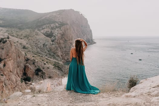 Side view a Young beautiful sensual woman in a mint long dress posing on a volcanic rock high above the sea during sunset. Girl on the nature on overcast sky background. Fashion photo