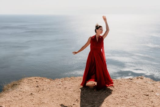 Side view a Young beautiful sensual woman in a red long dress posing on a rock high above the sea during sunrise. Girl on the nature on blue sky background. Fashion photo.