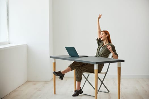 a very happy woman sits at a laptop and rejoices at the completed tasks raising her hands up. High quality photo