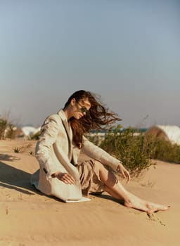 a woman in a stylish jacket and dark glasses sits on the sand relaxing. High quality photo