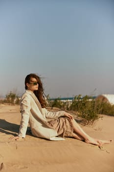 a woman in a stylish jacket and dark glasses sits on the sand relaxing. High quality photo
