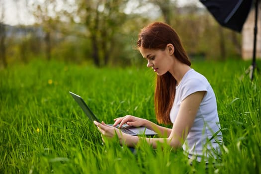 a woman in a light T-shirt sits in high green grass working at a laptop. High quality photo