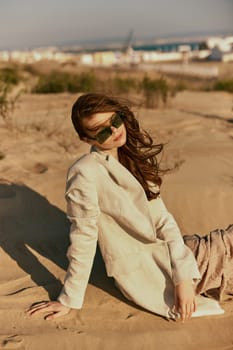 a woman in a stylish jacket and dark glasses sits on the sand relaxing. High quality photo