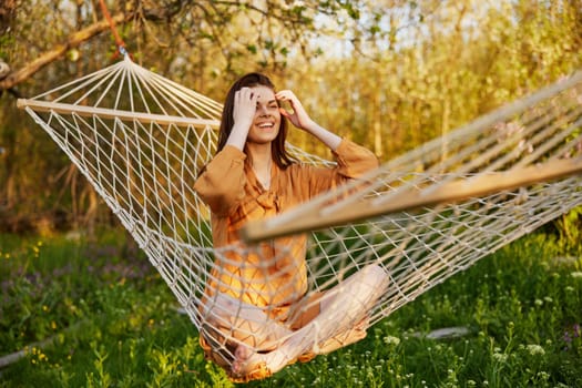 a happy woman in a long orange dress is resting lying in a hammock at the dacha, straightening her long hair and looking away, illuminated by the summer sun during sunset. High quality photo