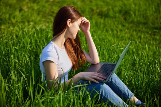 a woman works at a laptop sitting in a field covering her face from the sun with her hand. High quality photo