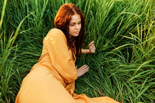 a happy woman is lying in the tall green grass in a long orange dress and smiling pleasantly looks away holding the leaves with her hand, enjoying nature. Close horizontal photo. High quality photo