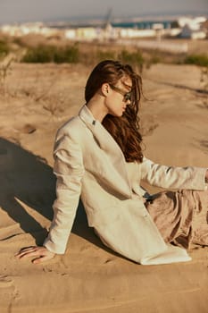 a woman in a stylish jacket and dark glasses sits on the sand relaxing. High quality photo