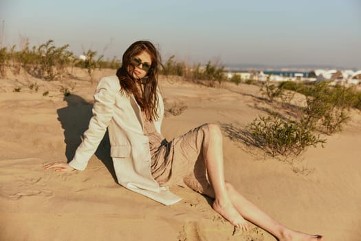 a woman in light clothes and black sunglasses sits on the sand in windy weather. High quality photo