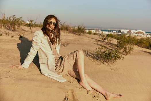 a woman in light clothes and black sunglasses sits on the sand in windy weather. High quality photo