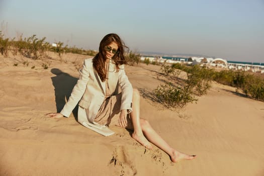 a woman in light clothes and black sunglasses sits on the sand in windy weather. High quality photo