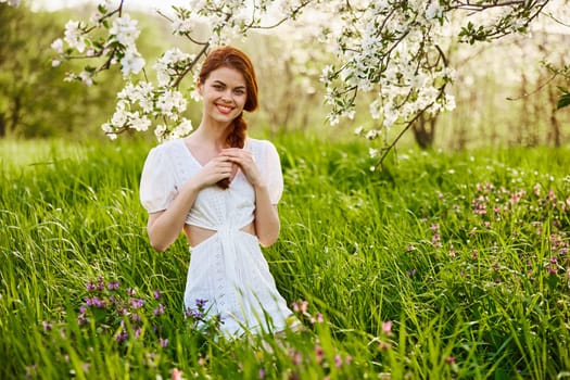 a young woman in a light summer dress stands near a tree with flowers and smiles looking at the camera. High quality photo