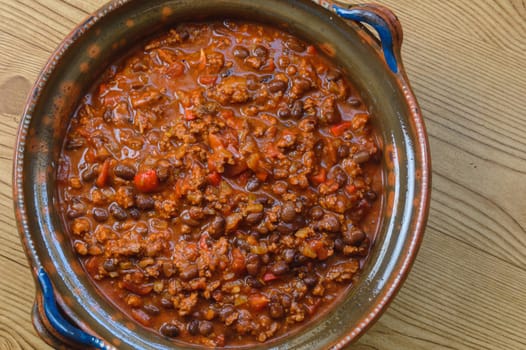 Black bean and ground beef chili in a large clay pot. Tex-Mex style chili dish. Homemade food. Red beef chili on wooden table top, flat lay shot. Top down view.