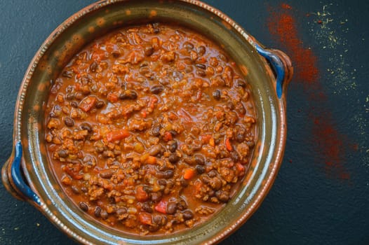 Black bean and ground beef chili in a large clay pot. Tex-Mex style chili dish. Homemade food. Red beef chili on black table top with red chili powder and Mexican oregano. Flat lay shot top down view.