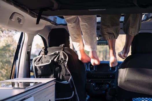 two unrecognizable women relaxing with feet dangling off the elevated bed of a camper van, concept of road trip adventure with best friend and van life relaxation