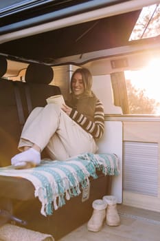 happy young woman relaxing reading a book in a cozy camper van at sunset, concept of road trip adventure and van life relaxation
