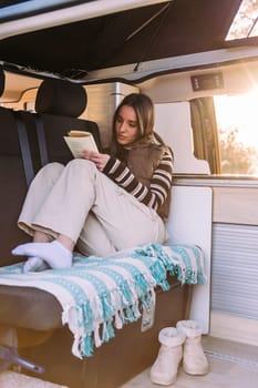 caucasian young woman relaxing reading a book in a cozy camper van at sunset, concept of road trip adventure and van life relaxation