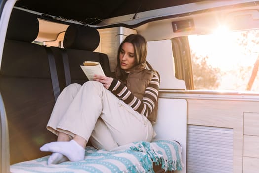 caucasian young woman relaxing reading a book in a cozy camper van at sunset, concept of road trip adventure and van life relaxation