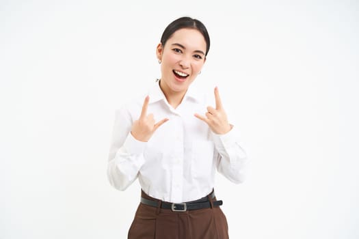 Excited smiling businesswoman, asian female employee, has fun, shows heavy metal, rock n roll gesture, celebrates, white background.