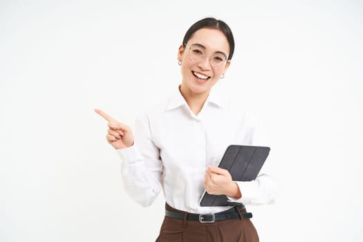 Young professional, businesswoman in glasses holding digital tablet, pointing finger left, showing business advertisement, standing over white background.
