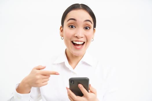 Close up portrait of happy saleswoman, successful woman with smartphone, pointing at her phone and smiling amazed, standing over white background.
