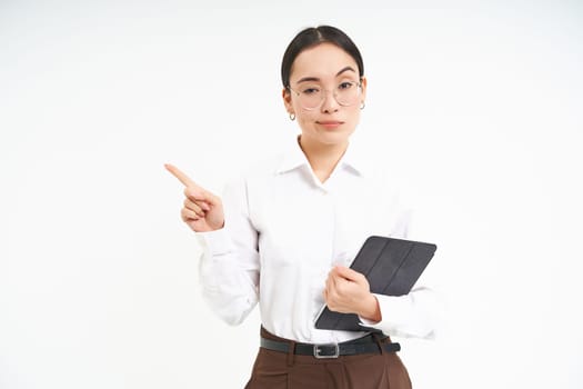 Portrait of korean corporate woman in glasses, holds digital tablet, points finger left and looks strict and serious, stands over white background.