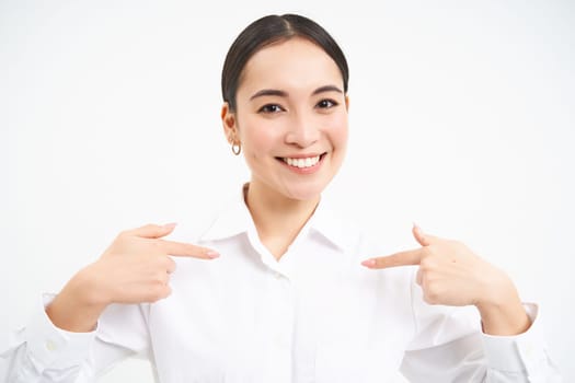 Asian businesswoman points at herself with confidence, looks like professional, standing isolated on white background.