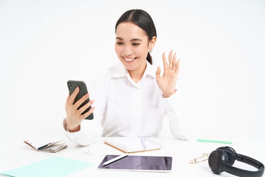 Young woman professional, businesswoman has a meeting on video chat, talks to mobile phone with client, stands over white background.