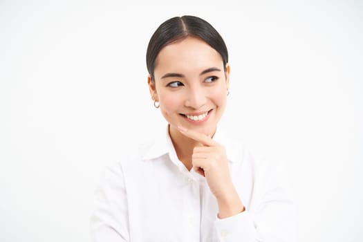 Successful asian woman, business professional, smiling with intrigued face, thinking, looking thoughtful aside, standing over white background.