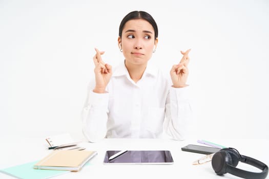 Hopeful asian businesswoman, cross fingers for good luck, sits in workplace office and makes wish, white background.