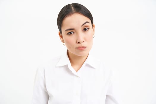 Portrait of skeptical asian woman, looks unamused and serious at camera, stands isolated on white background.