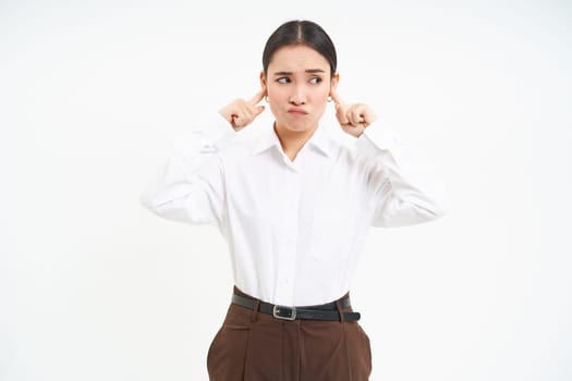 Frustrated young woman, shuts ears with fingers and grimaces from discomfort, annoyed by loud noises, white background.