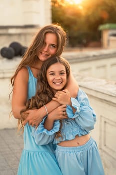 Portrait of mother and daughter in blue dresses with flowing long hair against the backdrop of sunset. The woman hugs and presses the girl to her. They are looking at the camera