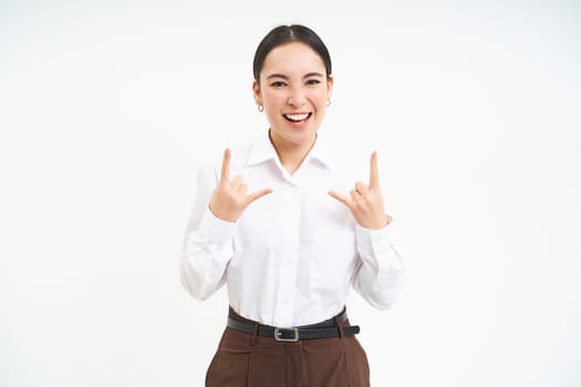 Excited smiling businesswoman, asian female employee, has fun, shows heavy metal, rock n roll gesture, celebrates, white background.