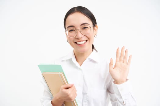 Image of friendly japanese student, girl waves hand, says hello, wears glasses, holds notebooks, stands over white background.