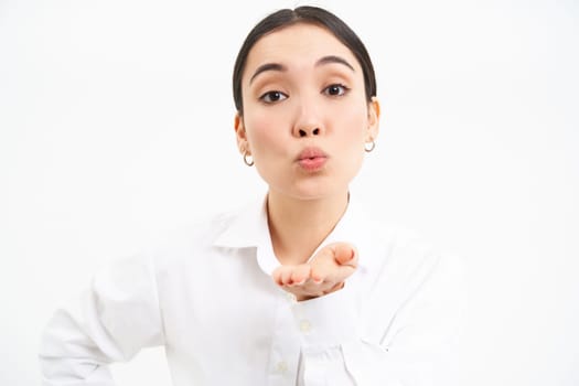 Close up portrait of cute young asian woman, blows air kiss with puckered lips, sends mwah at camera, standing over white background.
