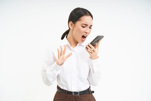 Angry business woman shouts at mobile phone, has intense conversation on telephone, stands over white background.