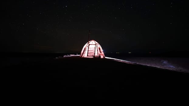 A tent with a stove stands under the starry sky. Night. Bright stars. Portable stove in the tent. The light is on. Lights are visible in the distance. Grass grows on the ground and snow lies in places