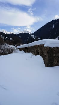 An old stone prison or hut in the mountains. Snowy mountains, forest and clouds against a blue sky. Tall dry bushes peek out from under the snow. Birch and spruce trees are visible in the distance.