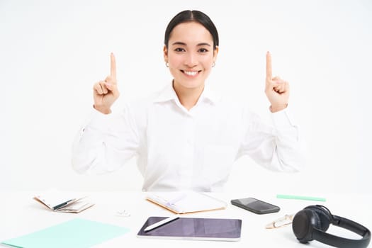 Business and profession. Portrait of asian woman in workplace, sits in office and points up, shows banner advertisement, white background.