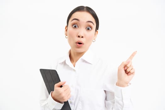 Image of young professional woman, pointing finger right, holding digital tablet, showing advertisement campaign, isolated on white background.