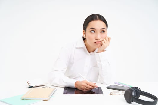 Tired and bored korean woman in office, employee sits with digital tablet, listens music in earphones and looks tired, white background.