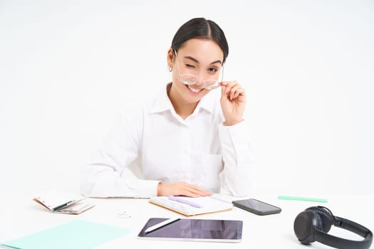 Portrait of successful businesswoman, office manager at workplace, sits with digital tablet and her working documents, wears glasses, smiles at camera.