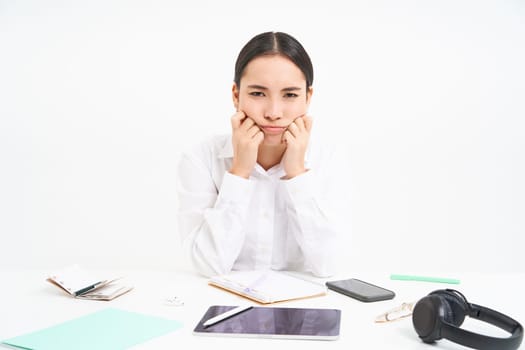 Sad and grumpy asian woman at workplace, sits with digital tablet and work documents, looks upset and bored, white background.