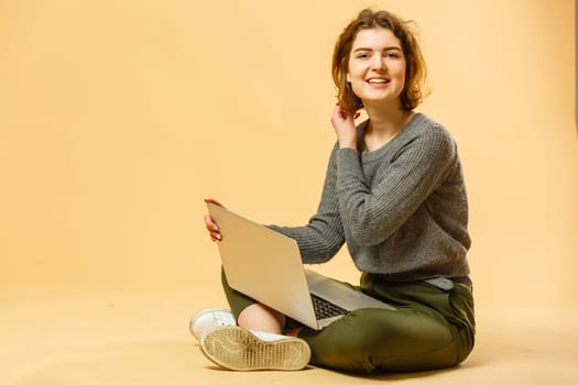 Happy young woman sitting on the floor with crossed legs and using laptop on beige background