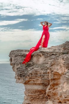 A woman in a red silk dress stands by the ocean, with mountains in the background, as her dress sways in the breeze