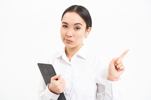 Portrait of serious asian woman with digital tablet, pointing finger right, showing banner, company advertisement, standing over white background.